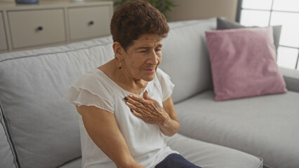 An elderly hispanic woman with short hair and mature features sits in a living room, clutching her chest in pain, depicting a health issue in a home interior setting.