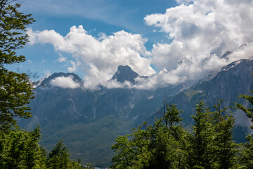 Obraz premium Scenic hiking trail through alpine pine forest. Majestic steep mountain peaks of massif Zhaborret shrouded in clouds, Albanian Alps (Accursed Mountains), Valbone Valley National Park, Albania. Hike