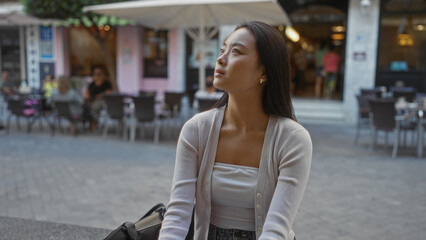 Obraz premium Asian woman sitting outdoors in an urban area, surrounded by people at a street cafe, looking pensive.