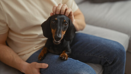 A man sits on a couch indoors with a dachshund dog on his lap, petting the dog's head affectionately in a cozy living room setting.