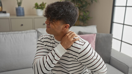 A young hispanic man in a striped shirt sits in a living room rubbing his neck, implying discomfort...