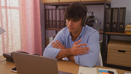 Handsome hispanic man working indoors, wearing headphones, using laptop in an office setting with...