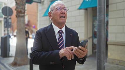 A senior caucasian man uses a smartphone on a city street while looking up, wearing a suit and striped tie, with urban buildings in the background.