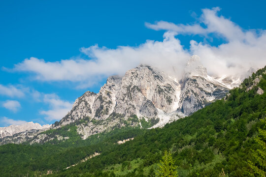 Majestic snow-capped mountain peaks of massif Zhaborret shrouded in clouds, Albanian Alps (Accursed Mountains), Valbone Valley National Park, Albania. Alpine slopes covered by lush green pine forest
