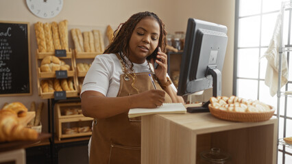 Plus size woman working in a bakery shop, taking notes, and talking on the phone while standing near a computer with a basket of pastries in the background