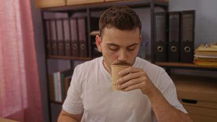 A young hispanic man with a beard is sipping a coffee in an office, surrounded by folders and boxes.