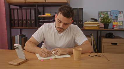 Handsome young hispanic man with a beard writing in a notebook in a workplace interior with office supplies, cup, and smartphone on a desk.