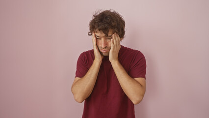 A distressed young hispanic man with a beard stands against a pink background, his hands on his...