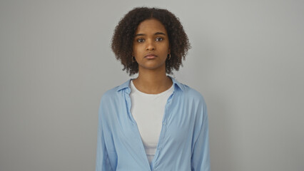A beautiful young african american woman with curly hair stands against an isolated white background, wearing casual clothes and looking at the camera with a calm expression. © Krakenimages.com