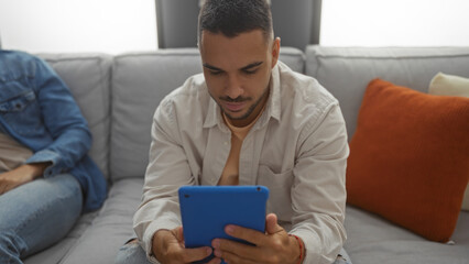 Man using tablet sitting on couch in living room with friend next to him, illustrating indoor...
