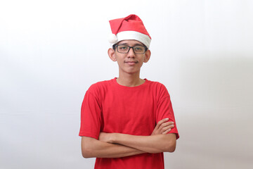 Portrait of excited Asian man in red t-shirt with santa hat keeping arms crossed and looking at camera. Isolated image on gray background