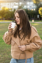 Woman in a beige coat holding a cup of coffee, gazing thoughtfully into the distance. Perfect for...