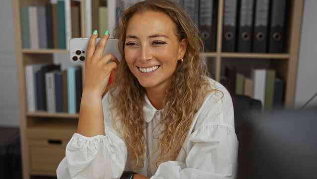 Blonde woman listening to a voice message on her smartphone while smiling in an office environment with books and folders in the background.