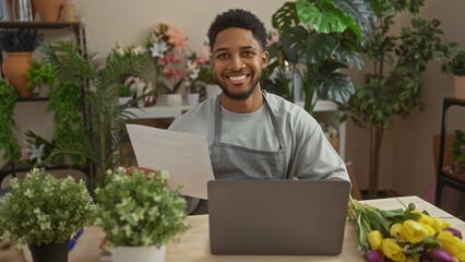 Smiling african man in apron reading at laptop surrounded by plants in flower shop