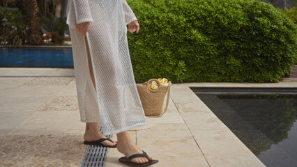 A woman walks by a pool at a bali resort, carrying a straw bag, embodying leisure and luxury.