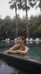 Fototapeta premium A smiling young woman in a swimsuit enjoys a leisurely day at a tropical balinese resort pool surrounded by palm trees.