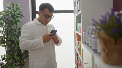 Fototapeta premium A young, handsome chinese man in a white coat uses his phone while standing in a pharmacy, surrounded by shelves of medical products and indoor plants.