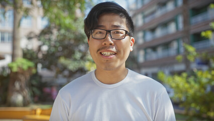 A smiling young asian man wearing glasses stands outdoors in a sunny city park.