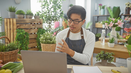 Handsome asian man in apron making video call at flower shop with plants in background
