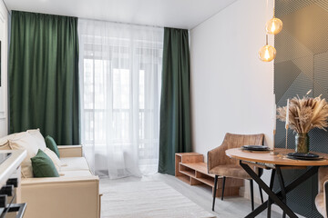 modern design of kitchen and living room. green curtains on windows. room has beige sofa, round table. romantic candlelit dinner is the table. kitchen is decorated in beige tones. Cozy apartments.