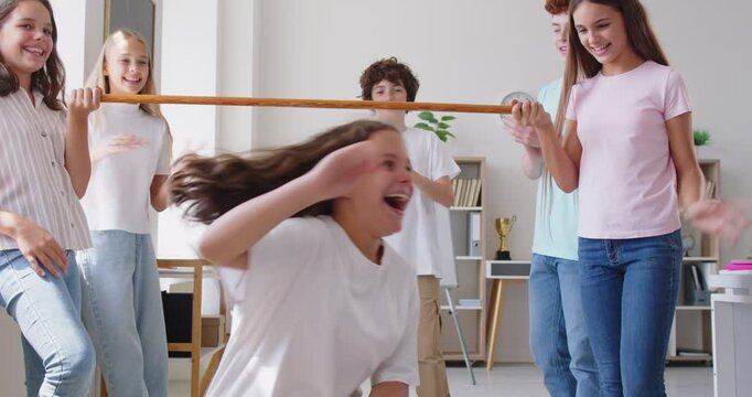 Cheerful caucasian high school children having fun together, taking part in limbo game and dancing during school party with friends. Group of happy classmates supporting participants with applause.