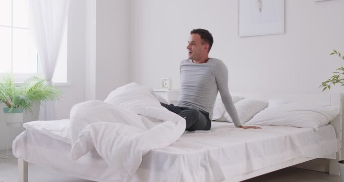 Man wakes up with smile on face and stretches out his hands, accepting positive energy of coming day. Cheerful young man gets up from white bed after nice nap and is ready to start his morning routine