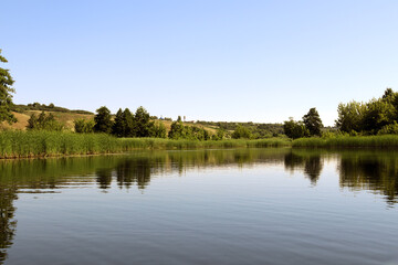 Beautiful serene river reflecting lush and trees greenery under clear blue sky at midday
