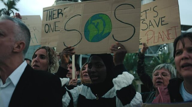Serious African woman activist marching holding banner in peaceful demonstration outdoor. Group diverse courageous people at manifestation on climate change and global warming pollution and factories