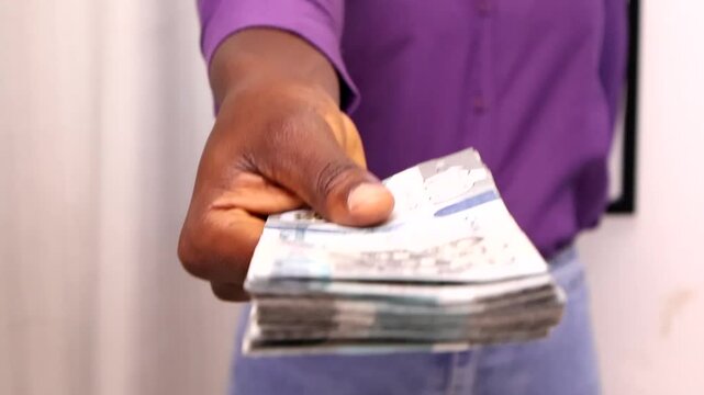 Young black african woman in purple long sleeves shirt and blue jeans throusers giving Ghanaian cedi notes to camera. paying Shopping payment purchasing