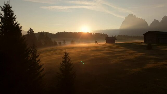 Drone shot capturing sunrise over misty meadows in Seiser Alm. Sunlight filters through trees, highlighting rustic cabins against the Dolomites mountains in the background.