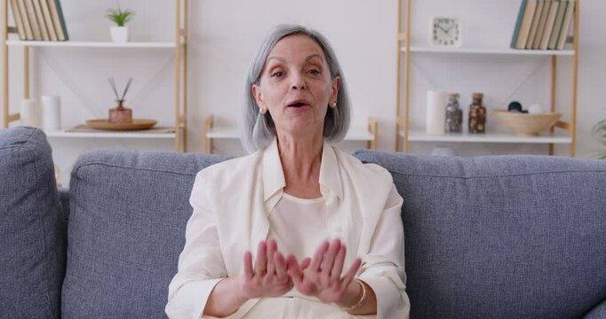 Happy old retired woman waving hand talking to webcam making online video call sitting on sofa at home. Senior female person chatting online with family. Concept of internet communication. 4k video.