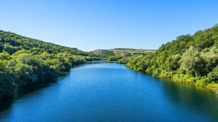 Obraz premium Serene River Landscape Surrounded by Lush Green Hills on a Clear Blue Sky Day
