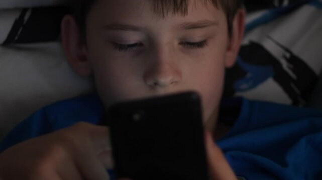 Caucasian boy 10 years old enthusiastically uses smartphone in the dark scrolls on the screen, teenager uses gadget lying in bed in the dark