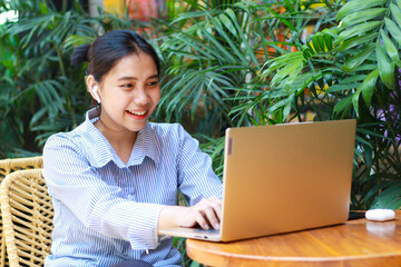 excited asian woman looking at laptop having video chat online meeting while sitting in cafe outdoor with green plant nature background wearing striped blue shirt