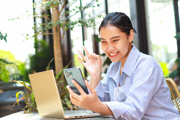 excited asian woman sitting in outdoor cafe with laptop on table holding smartphone showing ok...