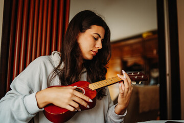 Young woman practicing ukulele guitar while reading notes on laptop	