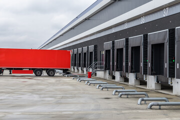 Red delivery truck at a modern distribution center with loading docks, roll-up doors, and a staircase entrance. Industrial warehouse scene for logistics and cargo handling.