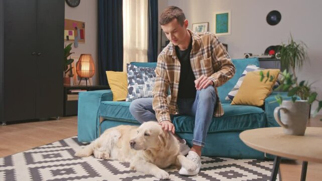 Retriever dog enjoying stroking hands of male owner while resting on floor at home. Young handsome man together with dog at modern peaceful apartment. Concept of pets, people and domestic animal.