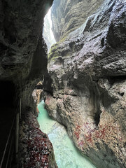 A narrow walkway winds through the rock cliffs at the valley's base alongside a flowing stream at Partnachklamm Gorge, Garmisch-Partenkirchen, offering a stunning alpine experience.