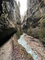 A scenic pathway follows the stream as it flows through the deep valley of Partnachklamm Gorge, surrounded by rocky cliffs and lush greenery in Garmisch-Partenkirchen, Germany.