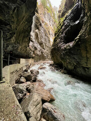 A narrow walkway winds through the rock cliffs at the valley's base alongside a flowing stream at Partnachklamm Gorge, Garmisch-Partenkirchen, offering a stunning alpine experience.
