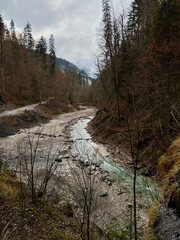 A stream flows from the valley of Partnachklamm Gorge into a lush forest with the majestic mountain range as a backdrop, creating a serene, natural scene in Garmisch-Partenkirchen.