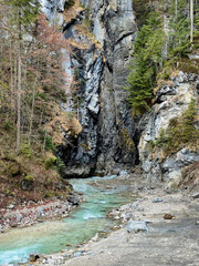 A crystal-clear blue river flows out from the rocky valley of Partnachklamm Gorge, framed by a dense forest and rugged mountain backdrop, creating a serene alpine scene.