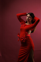 Fototapeta premium Beautiful young pregnant brunette girl. A woman waiting for a baby in a red dress with bows, on red background, in the studio.