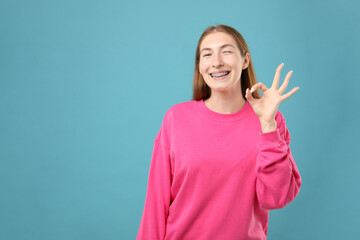 Smiling girl with braces showing ok gesture on light blue background, space for text