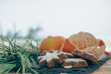 A cozy, festive arrangement showcasing traditional Christmas elements, including gingerbread star cookies, a rustic pine cone, fresh orange slices, and green pine branches. The setup evokes a warm hol