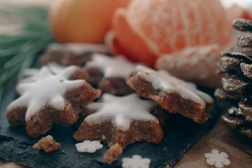 A cozy, festive arrangement showcasing traditional Christmas elements, including gingerbread star cookies, a rustic pine cone, fresh orange slices, and green pine branches. The setup evokes a warm hol