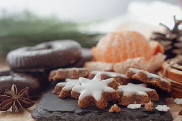 A cozy, festive arrangement showcasing traditional Christmas elements, including gingerbread star cookies, a rustic pine cone, fresh orange slices, and green pine branches. The setup evokes a warm hol