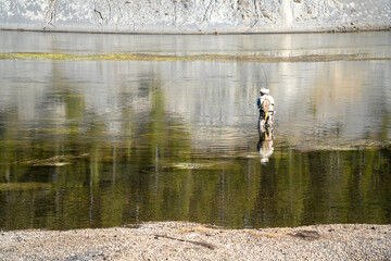 a fly fisherman in waders standing mid-river casting his line with a two-handed rod, trees reflecting in water