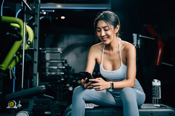 Asian woman resting on bench and checking all updates after exercising in gym interior, female using mobile phone application during training at fitness gym, sport workout healthy lifestyle concept